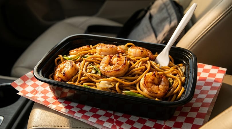 Shrimp lo mein in a black takeout container with a white fork, resting on a red and white checkered paper in a car seat.