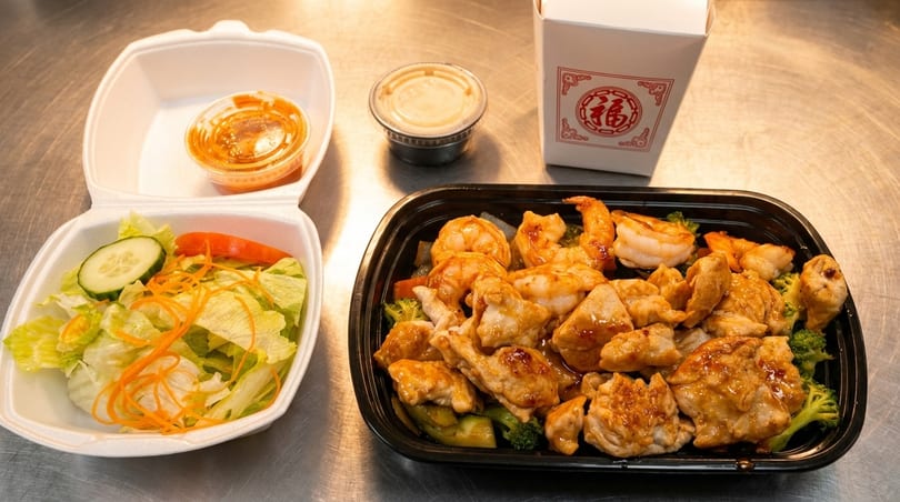 Chinese takeout meal with chicken and shrimp, a side salad, and sauces on a metal surface.